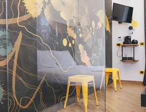 A modern room with a glass partition, a grey sofa, two yellow stools, a flat-screen TV, and a coffee machine on a shelf. - Amarillo B&B (Gaeta)