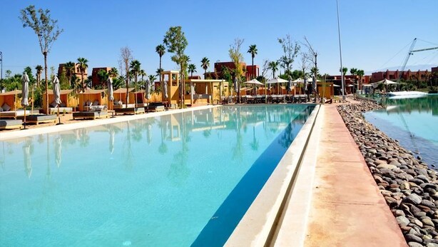 A clear swimming pool with lounge chairs and umbrellas, surrounded by palm trees and buildings. - Villa Jasmine V4 70 Marrakesh (Marrakech)