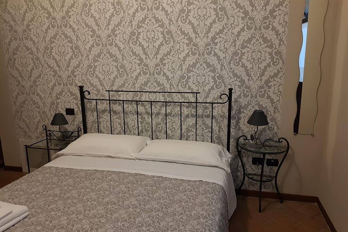 Un lit bien fait, avec une couvre-lit à motifs, une tête de lit en métal, deux tables de chevet avec des lampes et un papier peint à motifs.