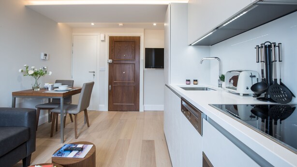 Ett modernt kök med en inbyggd diskmaskin, en matplats med ett träbord och stolar samt en liten sittgrupp med en soffa och ett soffbord.