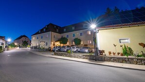 A street view at dusk with a row of buildings, including a hotel with a sign indicating it is a restaurant. There are parked cars and a street lamp. - Landhaus Krone (Steinach)