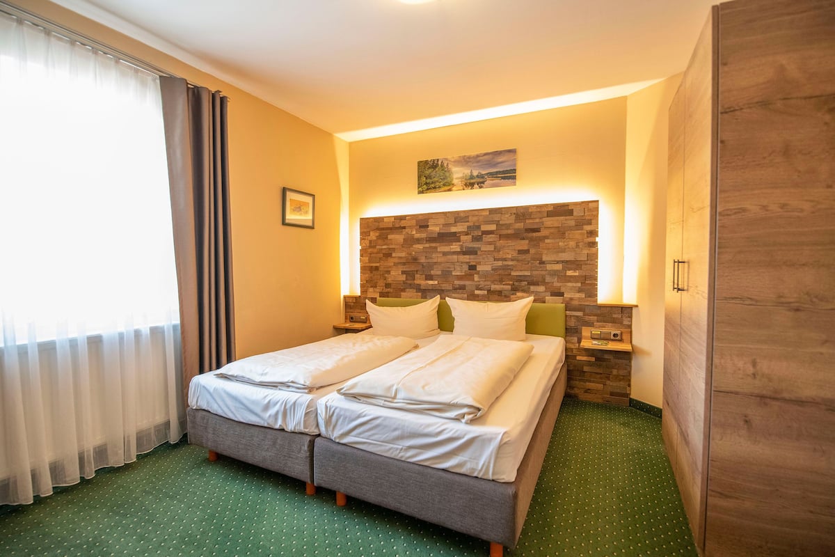 A double bed with a wooden headboard, two bedside tables with lamps, a green carpet, and a window with curtains.