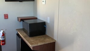 A small hotel room with a bed, a desk with a flat-screen TV, a small kitchenette, and a fire extinguisher. - Karbani Inn (Carlsbad)