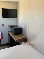 Una habitación de hotel pequeña con cama, escritorio con televisor de pantalla plana, kitchenette y extintor.