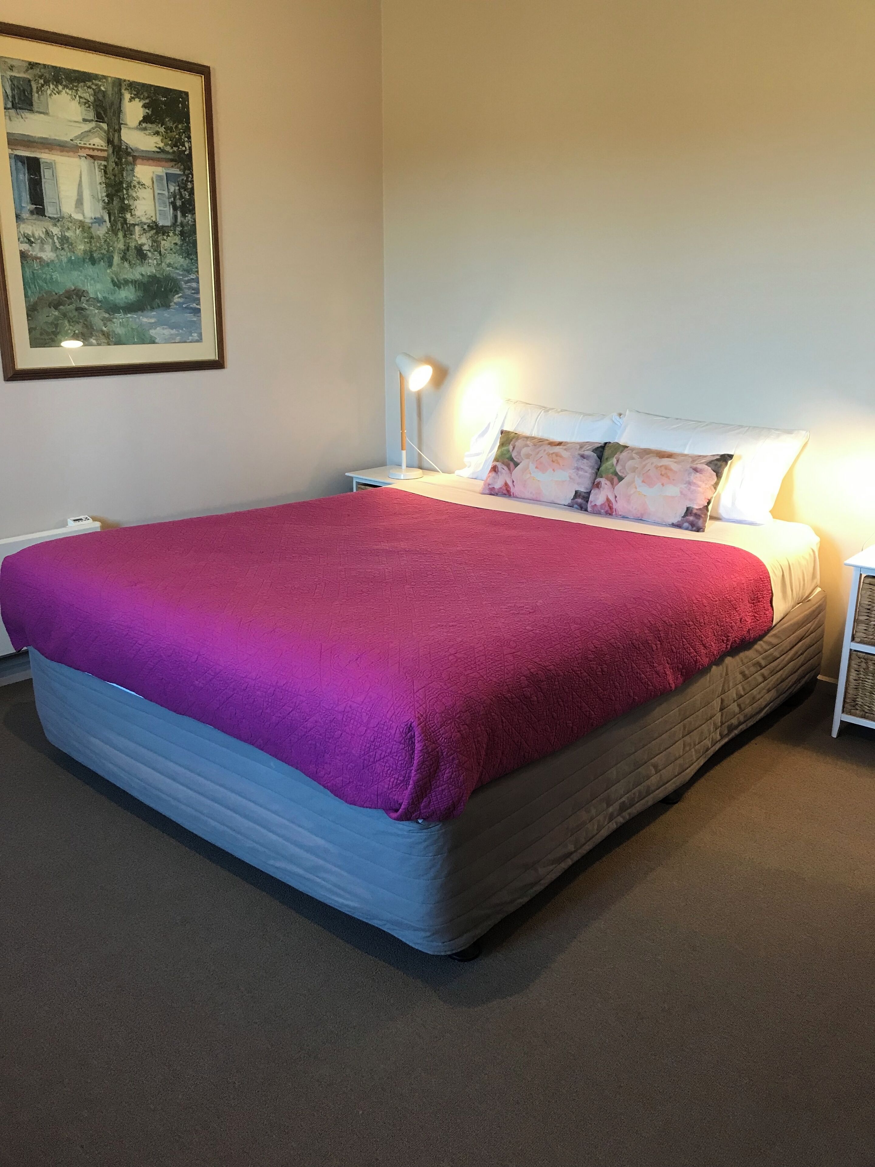 A neatly made bed with a red blanket, a white pillow with a floral design, and a bedside lamp on a nightstand.