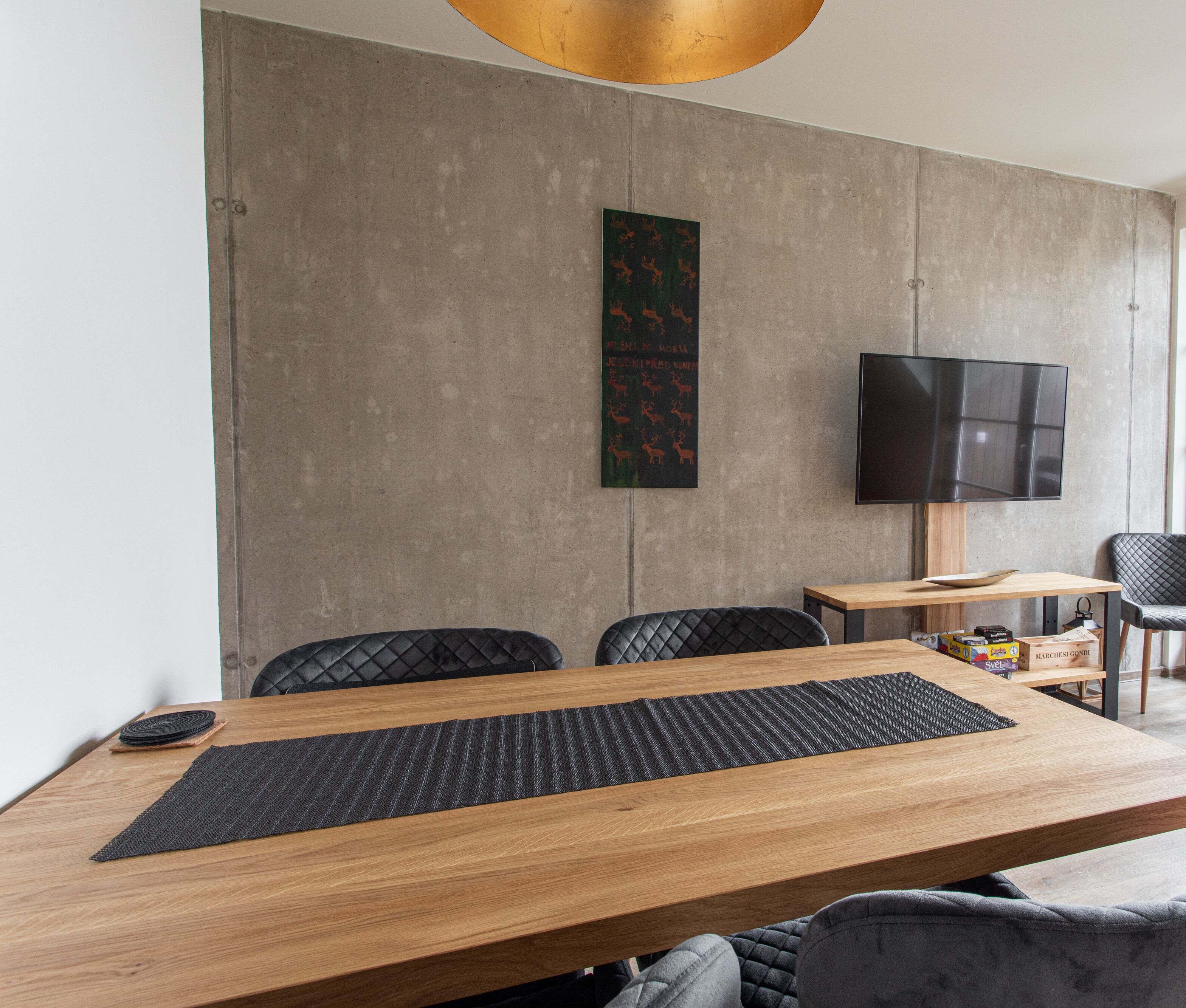 A modern dining area with a wooden table, black chairs, a black woven placemat, a mounted flat-screen TV, and a decorative wall piece.