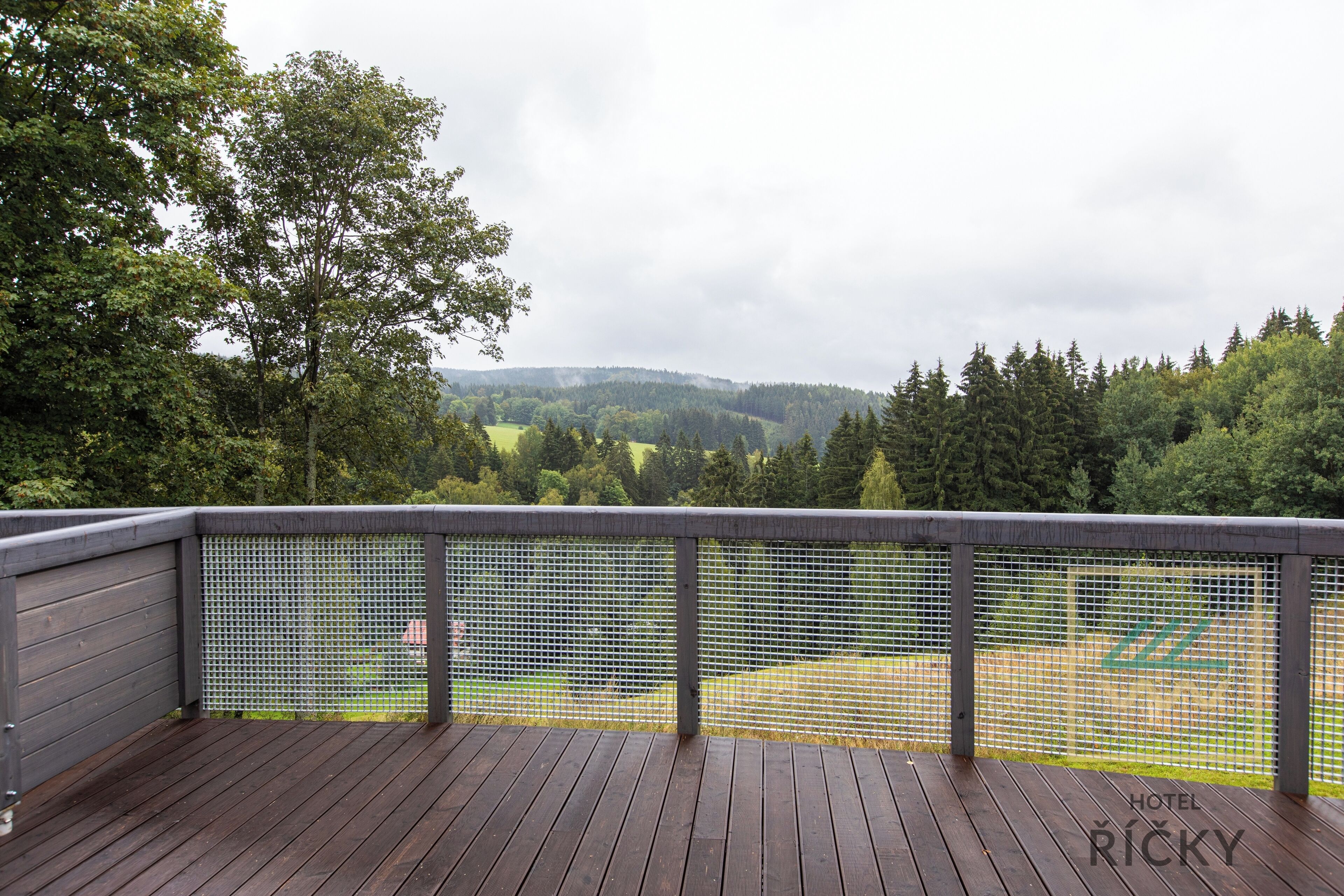 A wooden deck with a mesh railing overlooking a green landscape and a hilly area.