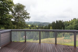 A wooden deck with a mesh railing overlooking a green landscape and a hilly area.