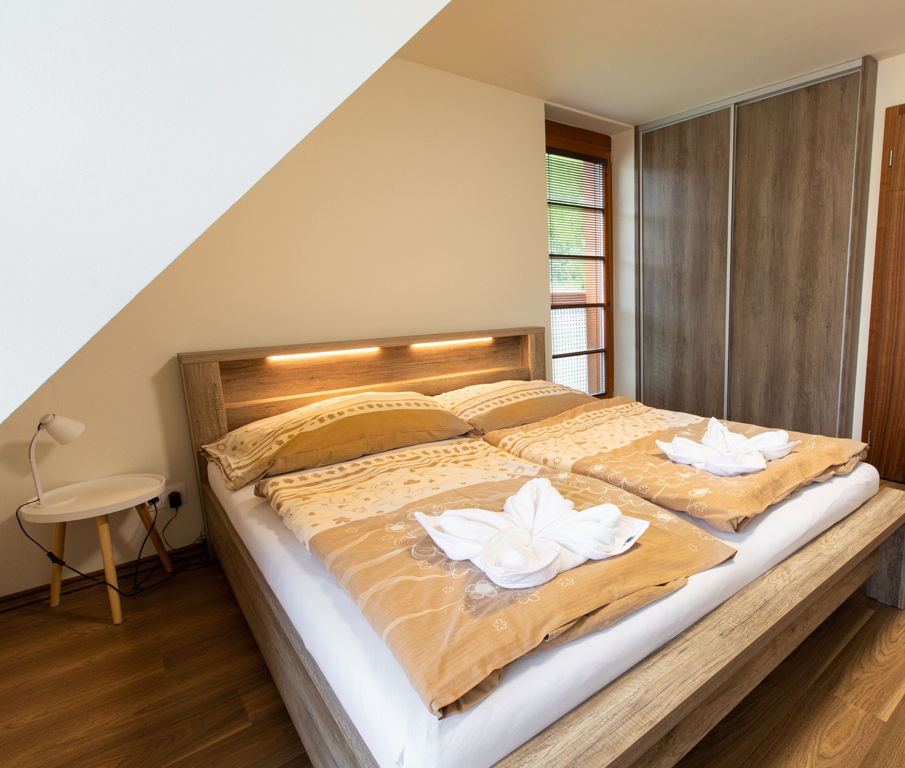 Un dormitorio con cabecera de madera, cama con ropa de cama color beige, lámpara de noche y ventana con persianas.