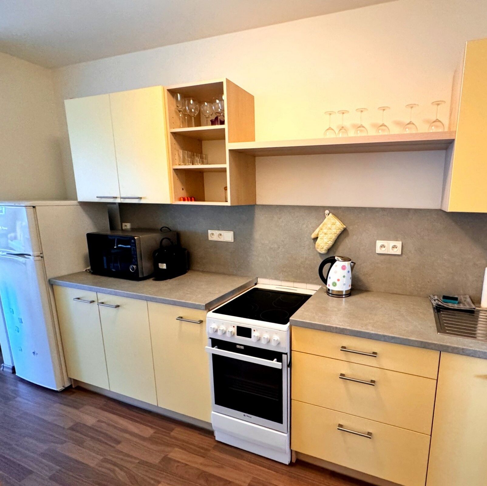 A compact kitchen with a stove, oven, and a microwave. There are cabinets above and below the counter, and a shelf with glasses.