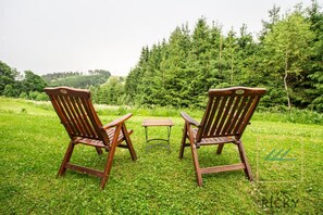 Two wooden Adirondack chairs and a small table set on a grassy lawn with a forest in the background.