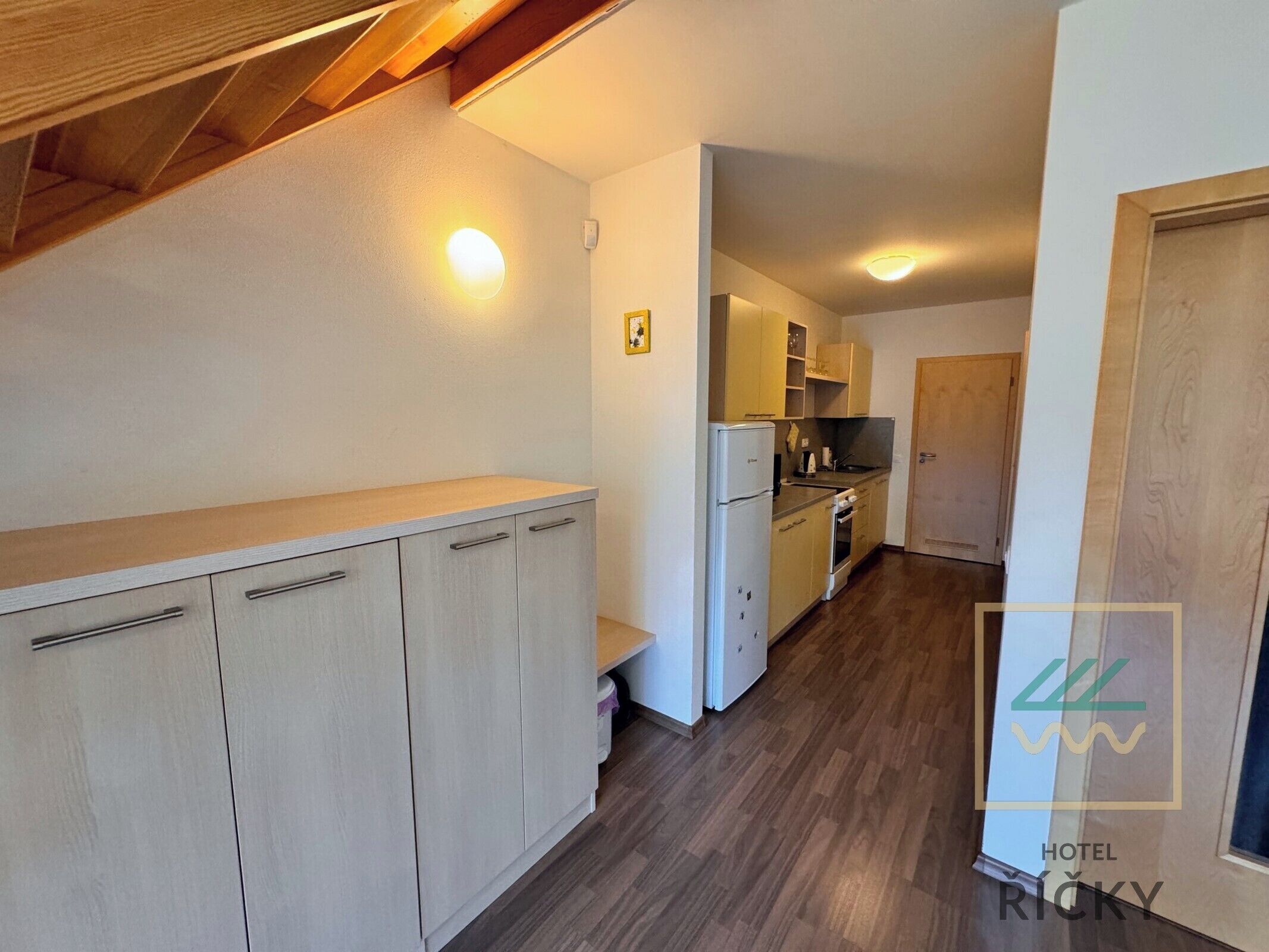 A narrow hallway with a kitchenette on the right, wooden flooring, and a door at the end.