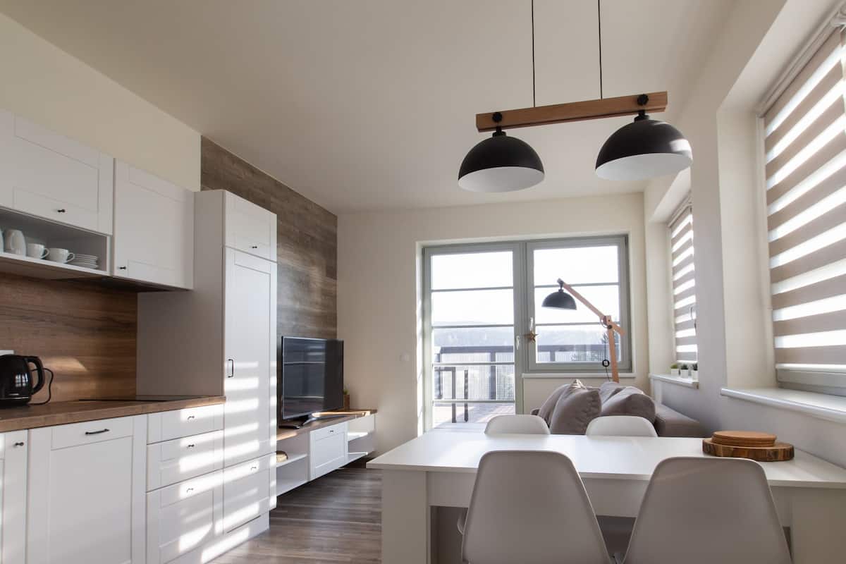 A modern kitchen with white cabinets, a wooden accent wall, a dining area with a white table and chairs, and a large window with blinds.