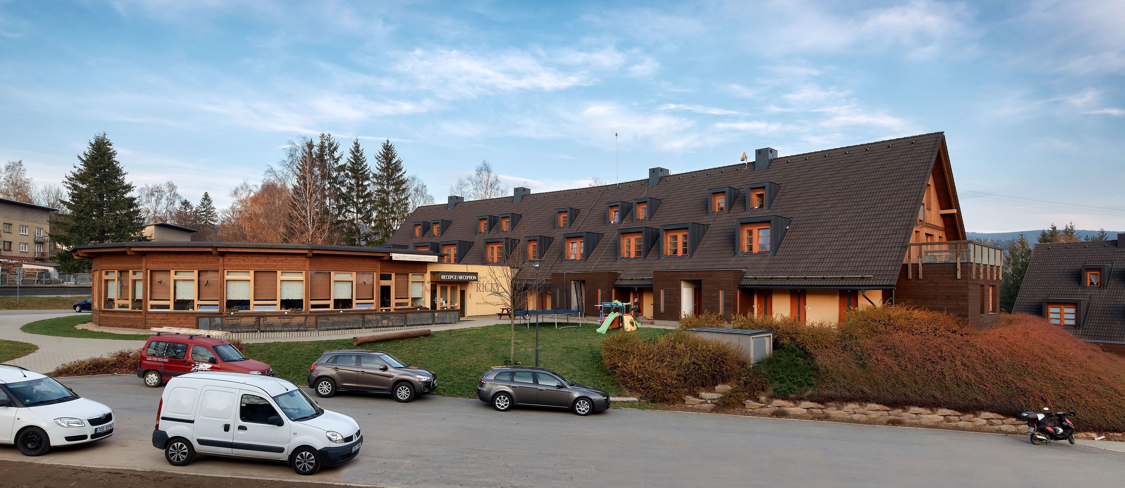 A modern building with multiple windows and a sloped roof, surrounded by parked cars and a small playground.