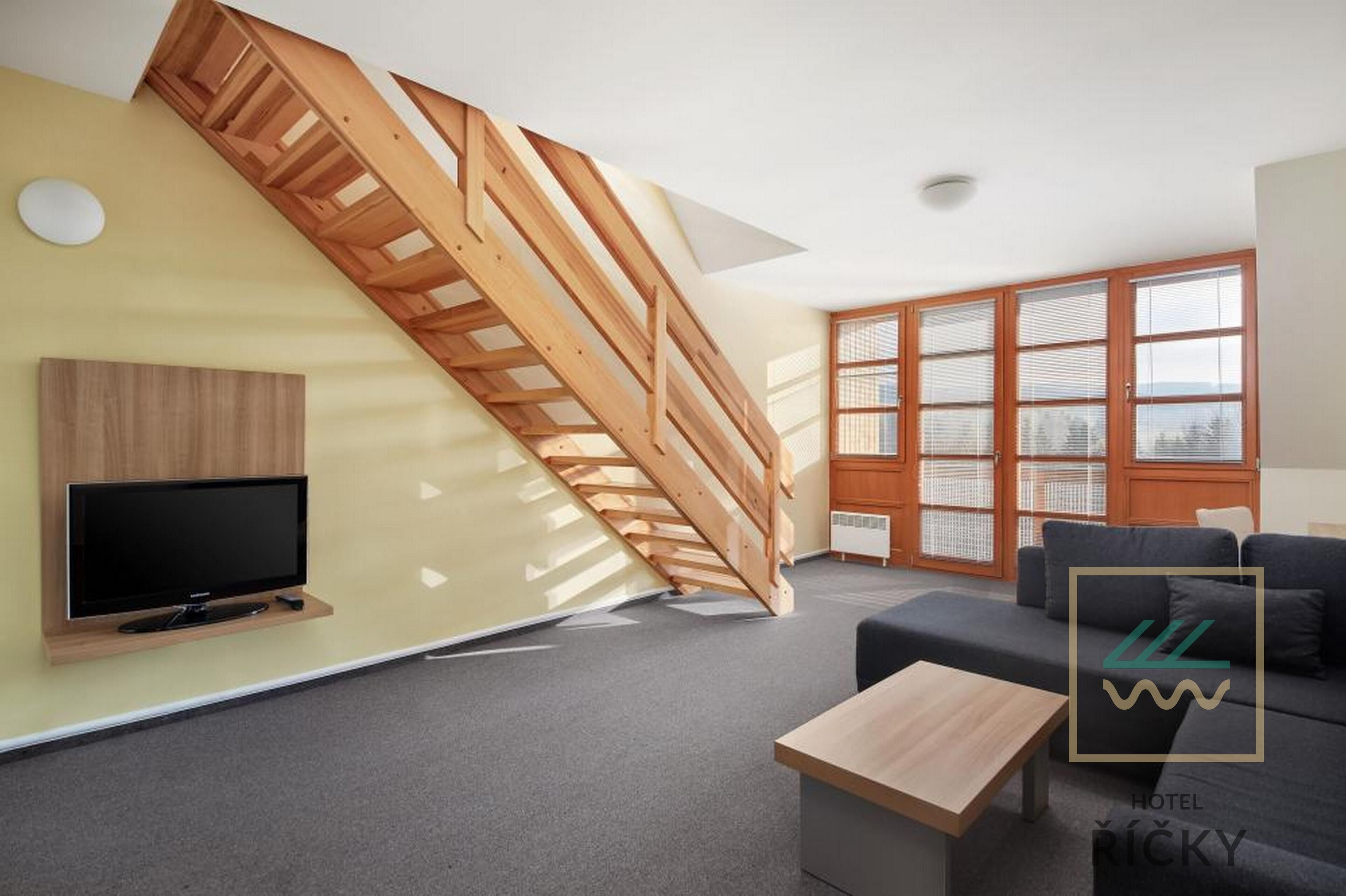 A modern hotel room with a wooden staircase, a flat-screen TV, a sofa, and a large window with blinds.