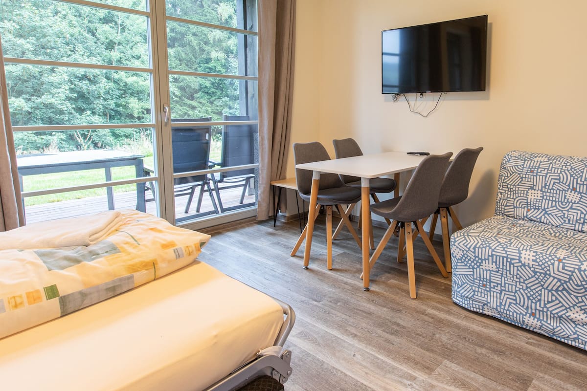 A hotel room with a bed, a sofa, a dining table with chairs, a TV mounted on the wall, and a large window with a view of trees.