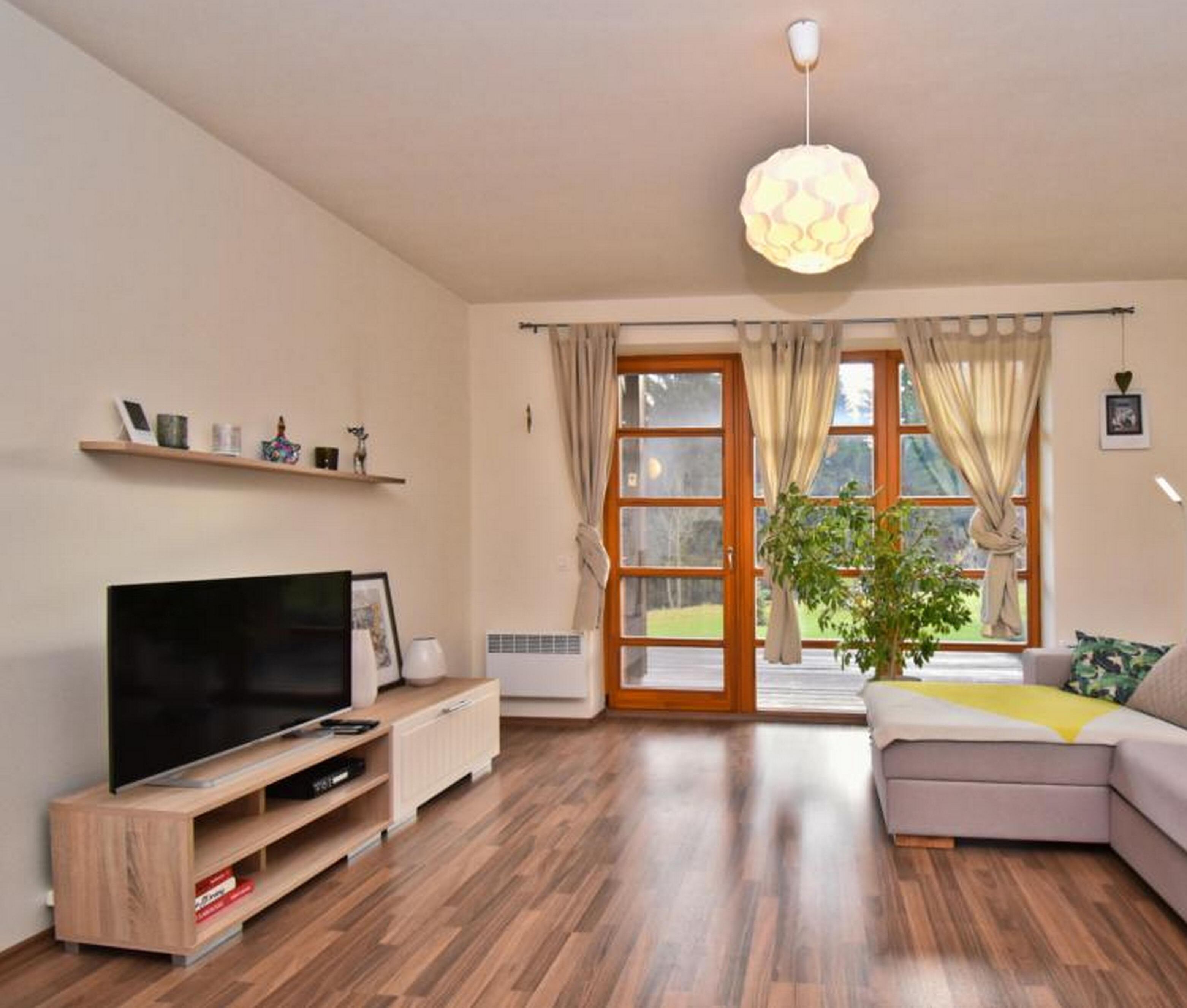 Een woonkamer met een flatscreen televisie, houten vloer, een bank, een plant en een groot raam met gordijnen.