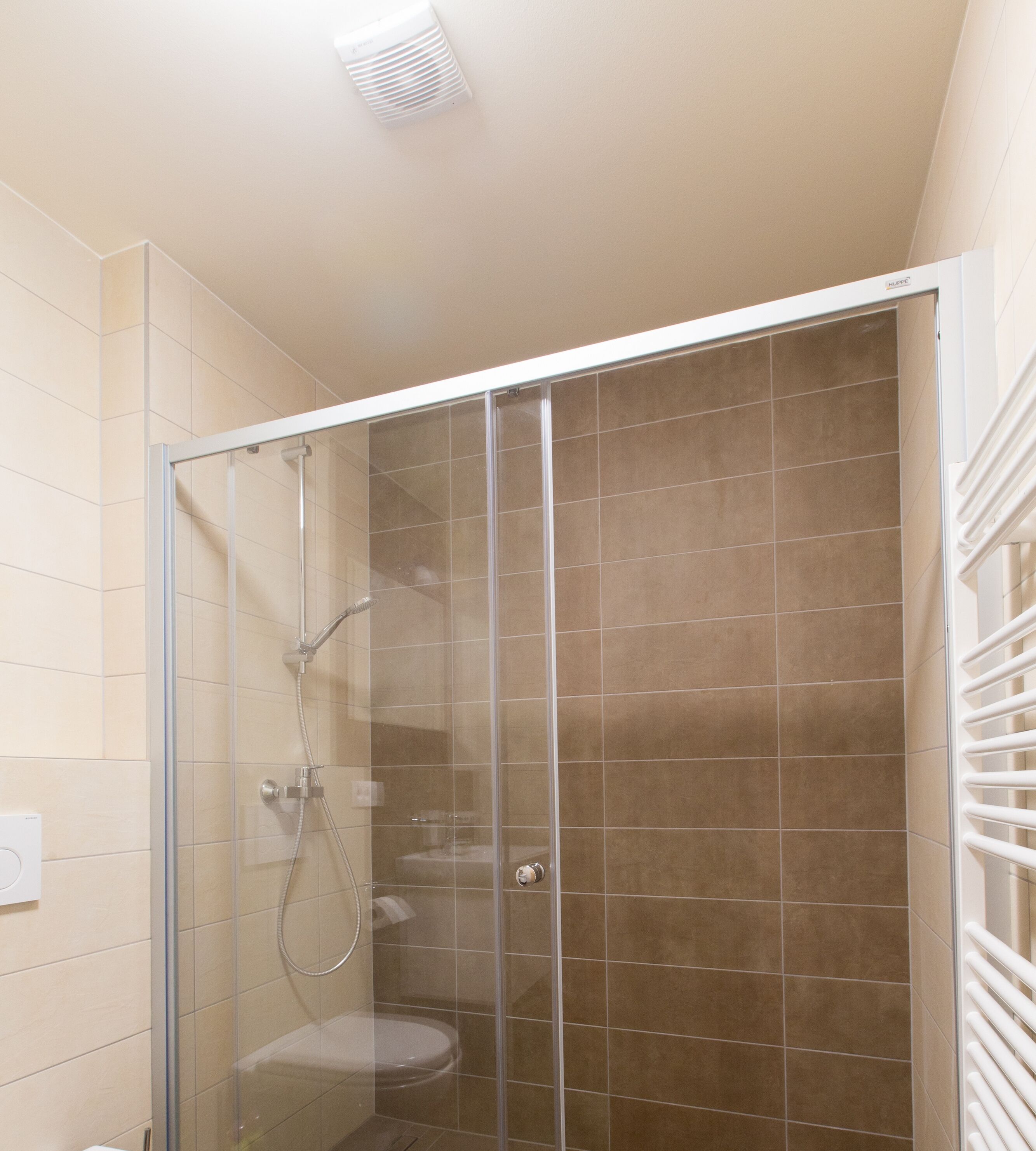 A glass-enclosed shower with a white ceiling light, beige tiles, and a white towel rack.