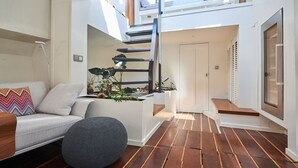 A modern interior with a staircase leading to a loft, a grey sofa, a round ottoman, and wooden flooring.