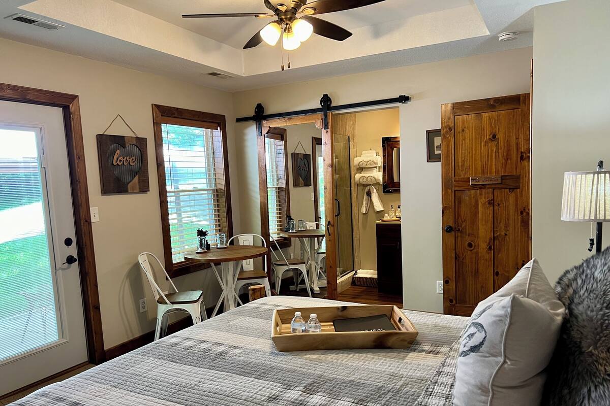 A bedroom with a bed, a wooden nightstand, a desk with chairs, a shower area, and a door with a towel rack.