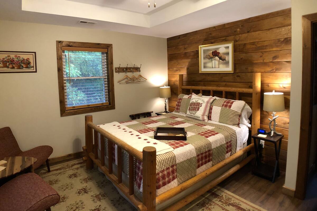 A cozy bedroom with a wooden headboard, a bed with a quilt, a bedside table with a lamp, a chair, and a window with blinds.