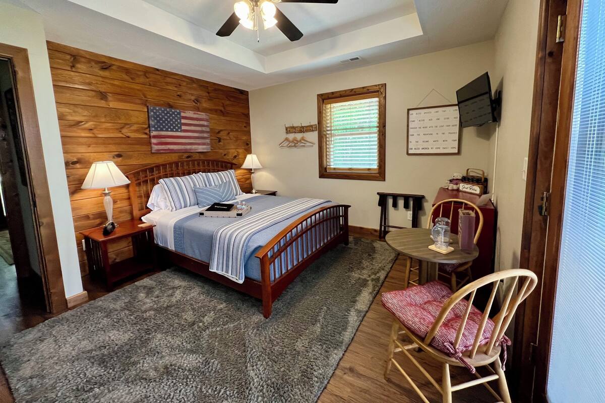 A bedroom with a wooden headboard, a bed with a blue and white striped bedspread, two bedside lamps, a ceiling fan, a small table with a chair, a wooden chair with a red and white checkered cushion, a window with blinds, and an American flag on the wall.