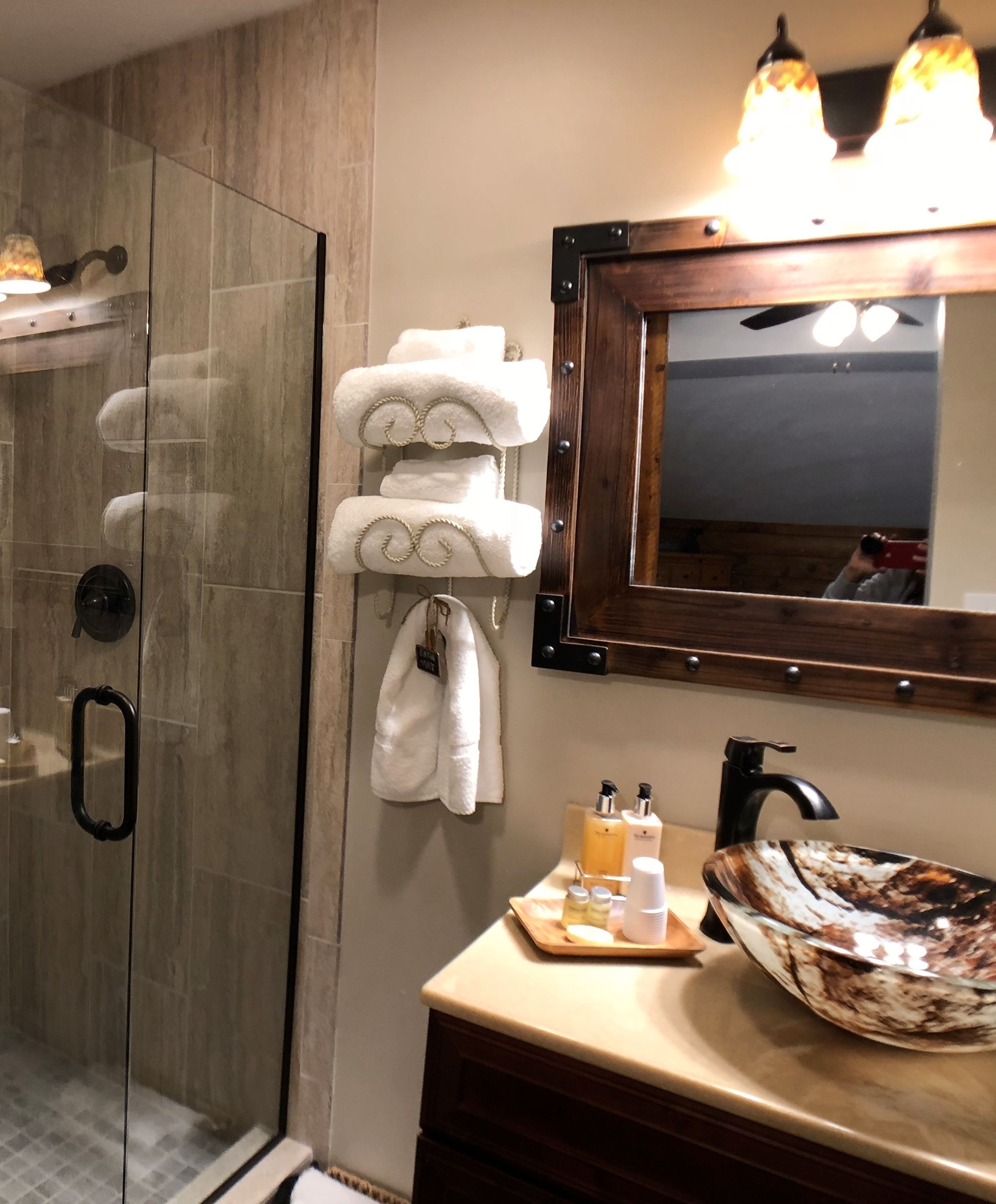 Un baño con cabina de ducha de vidrio, tocador de madera con un lavamanos grande y un espejo con marco de madera.