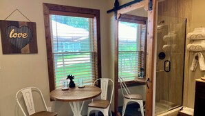 A bathroom with a glass shower, white chairs, a small round table, and a window with blinds. - Walnut Waters Bed & Breakfast (Lone Jack)