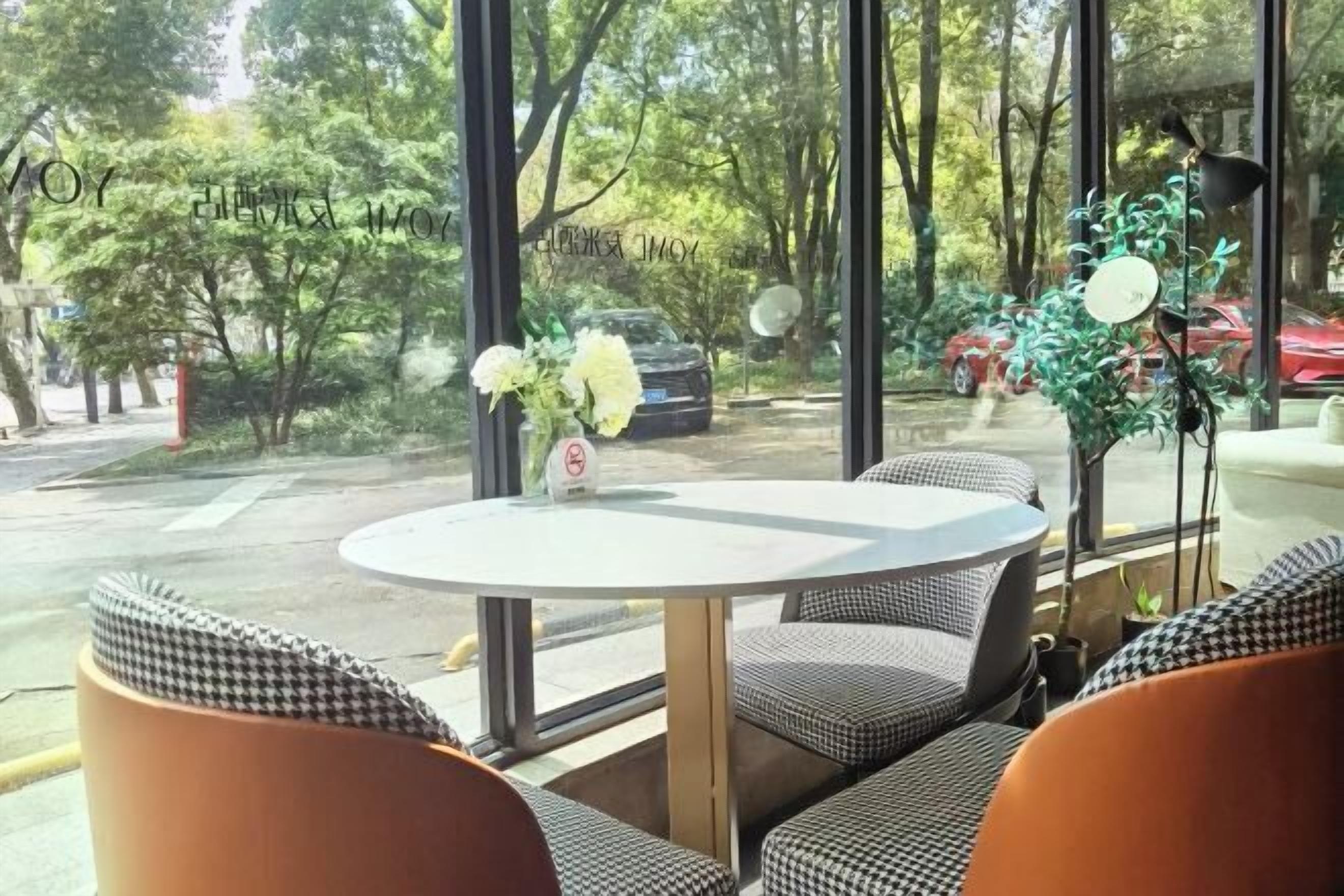 A round table with checkered cushions, a vase with flowers, and a view of a parking area with trees and cars.