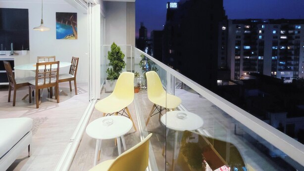 Eine moderne Wohnung mit Balkon, ausgestattet mit gelben Stühlen und einem Glastisch, einem Essbereich mit Holzmöbeln und Blick auf die Stadt in der Abenddämmerung.