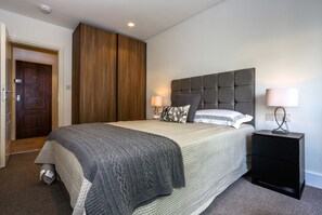 A bedroom with a large bed, a wooden wardrobe, a nightstand with a lamp, and a door. - London Villa - 20 Princes Square (London)