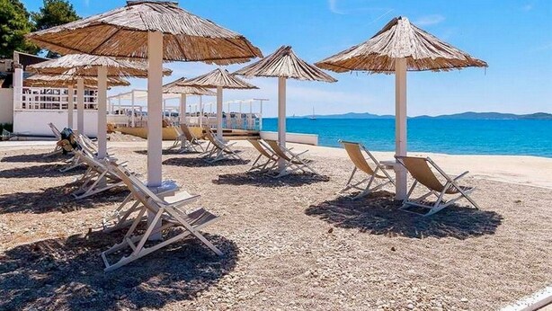 Une plage avec des chaises longues et des parasols en chaume, un ciel bleu dégagé et une mer calme.