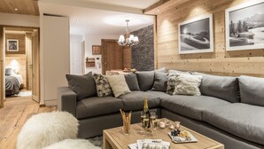 A modern living room with a sectional sofa, wooden flooring, and a chandelier. There are framed pictures on the wall and a coffee table with snacks and drinks. - Whistler Lodge Apartment T3 Cabin B07 (Courchevel)
