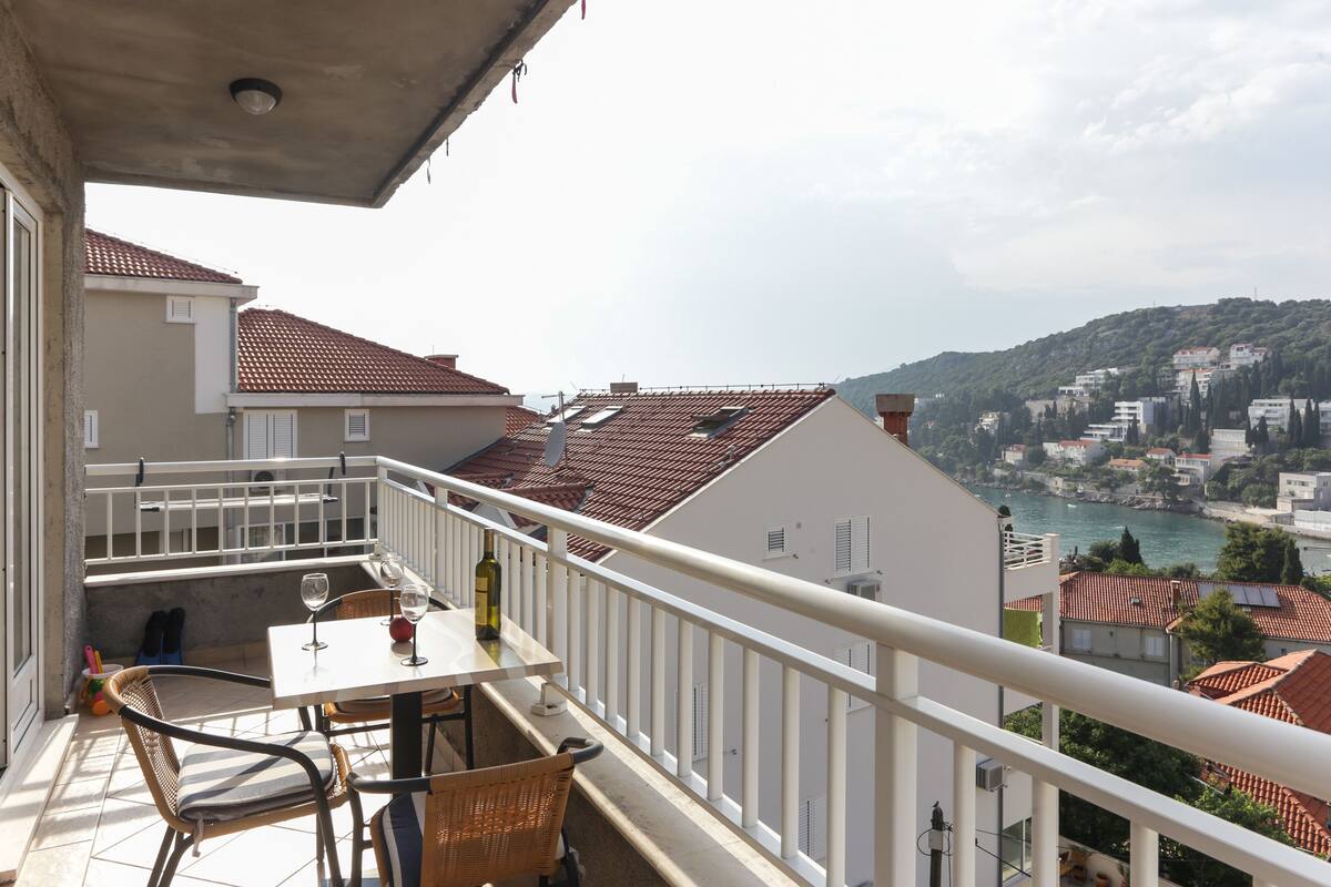 A balcony with a table and chairs, overlooking a coastal town with terracotta roofs and a body of water.