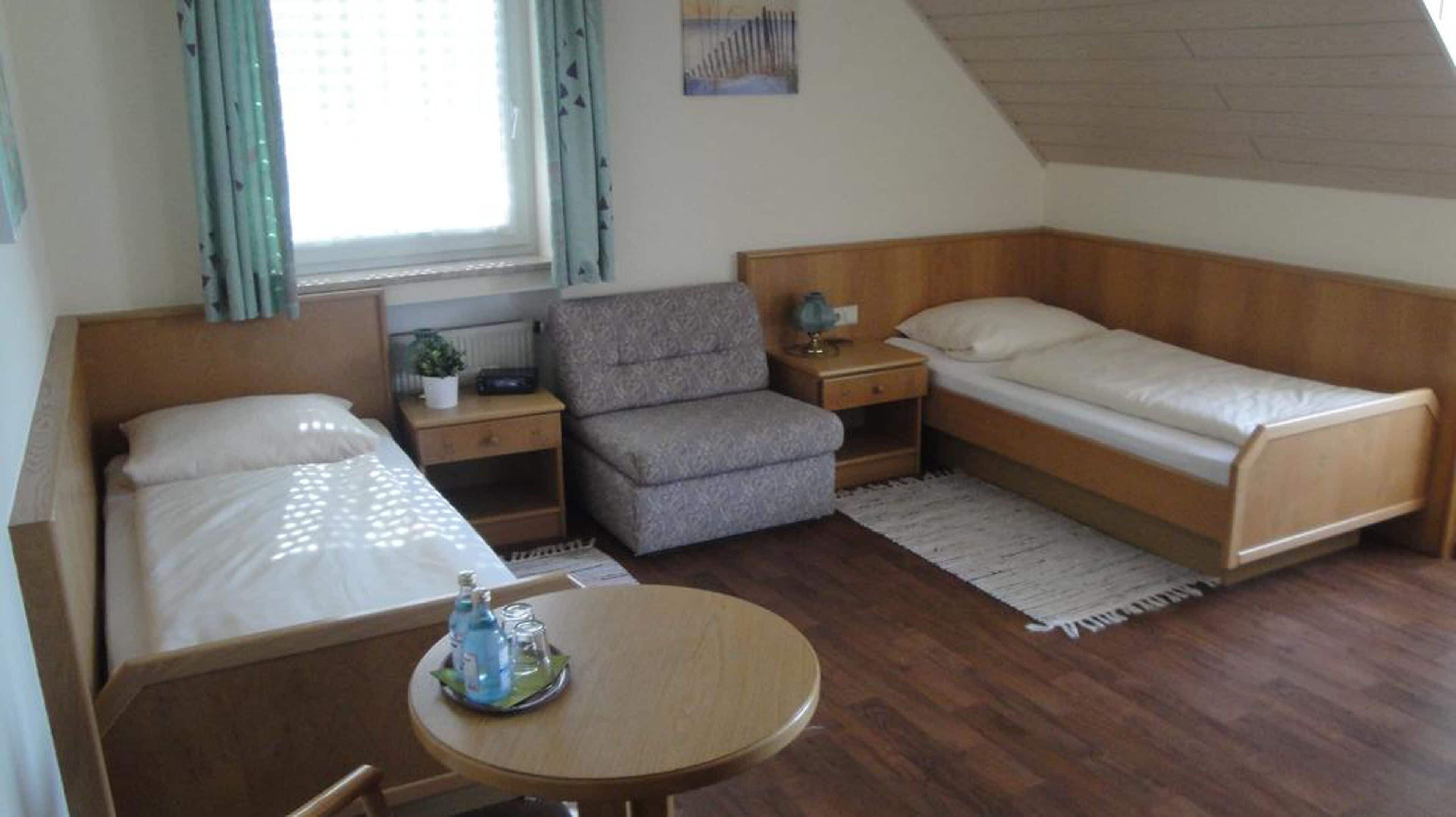 Una habitación pequeña y luminosa con dos camas individuales, un sofá, una mesita con una botella y vasos, y una ventana con cortinas.