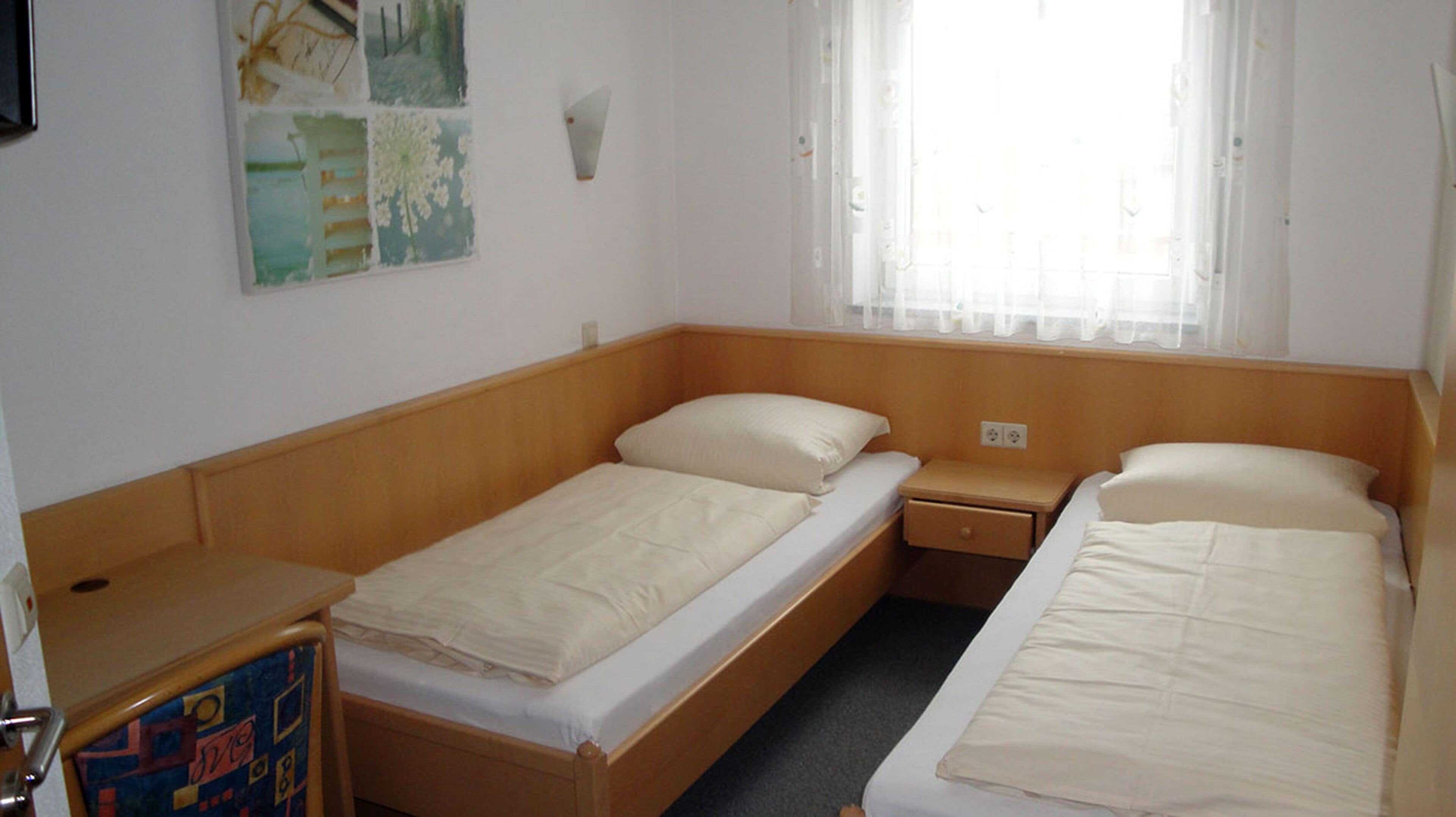Ein kleines Hotelzimmer mit einem Einzelbett, zwei Betten, einem Holzkopfende, einem kleinen Nachttisch und einem Fenster mit durchsichtigen Vorhängen.
