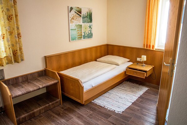 Una habitación pequeña y ordenada con una cama de madera, una mesita de noche, un pequeño banco y un cuadro colgado en la pared.
