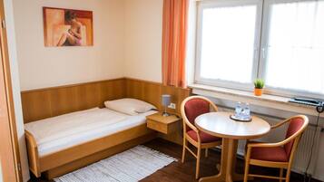 Ein kleines Hotelzimmer mit einem Einzelbett, einem Tisch mit zwei Stühlen, einem Fenster mit Vorhängen und einem Bild an der Wand.