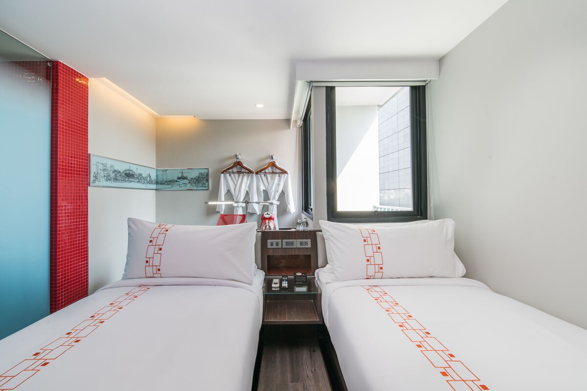 Une chambre d’hôtel avec deux lits, une table de chevet, une fenêtre et un mur recouvert de carreaux rouges.