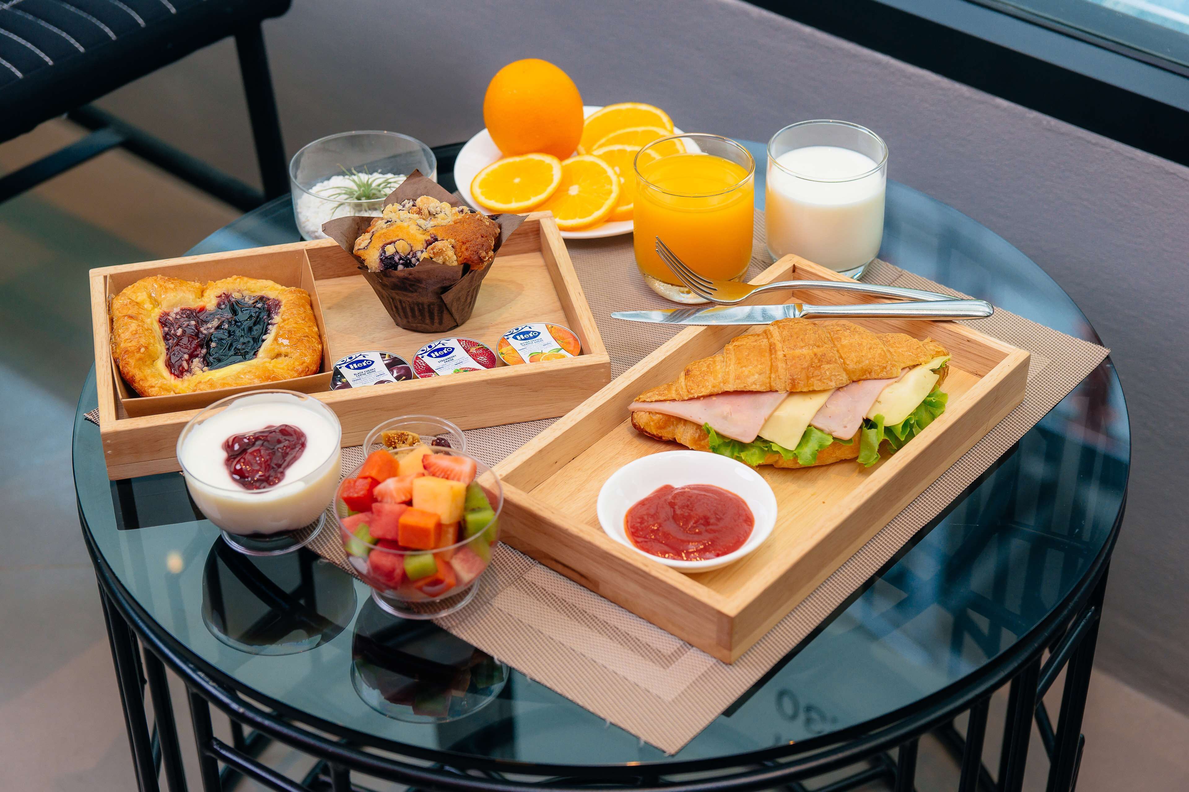 A breakfast tray with a croissant, muffin, fruit salad, and a sandwich on a glass table.