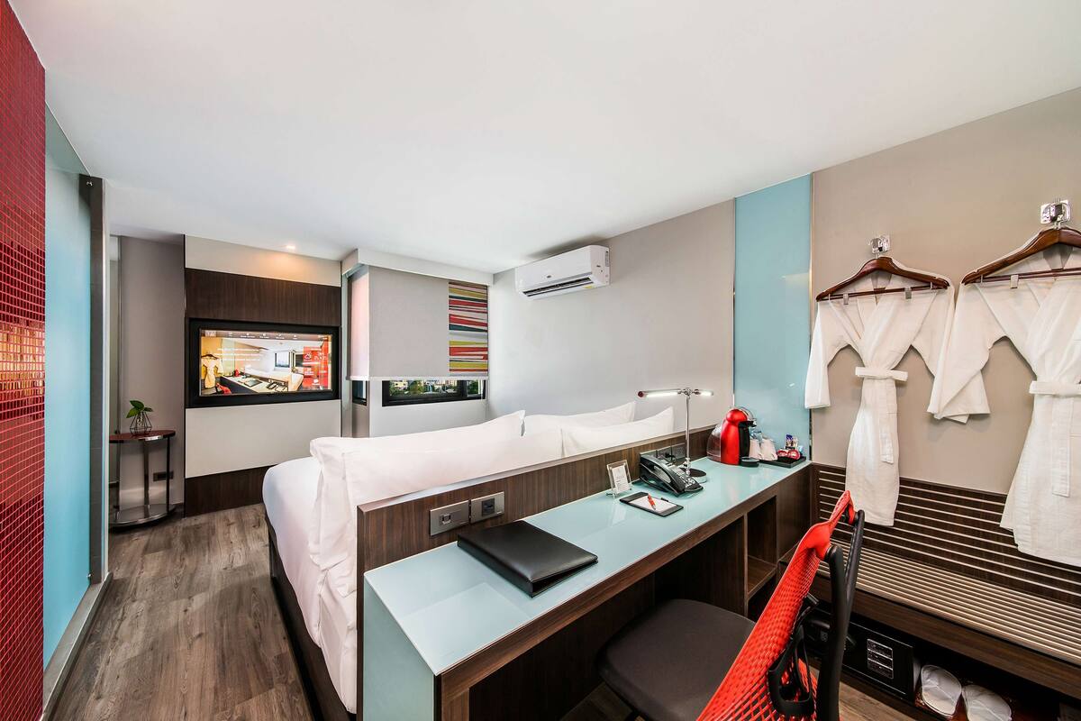 Une chambre d’hôtel avec un lit, un bureau et un coin salon. On y trouve une télévision, une lampe de bureau et une chaise rouge.