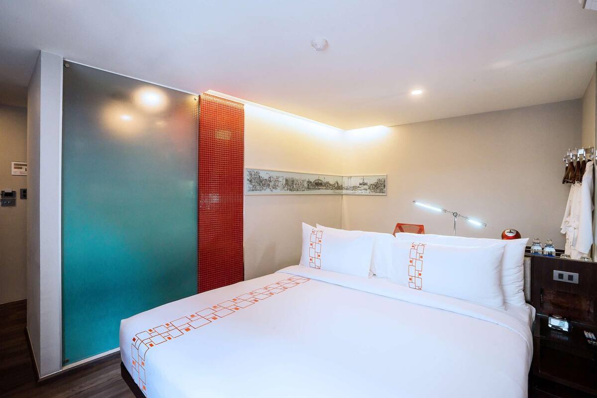 Une chambre d’hôtel avec un lit, une table de chevet, une commode, une lampe fixée au mur et un paravent rouge.