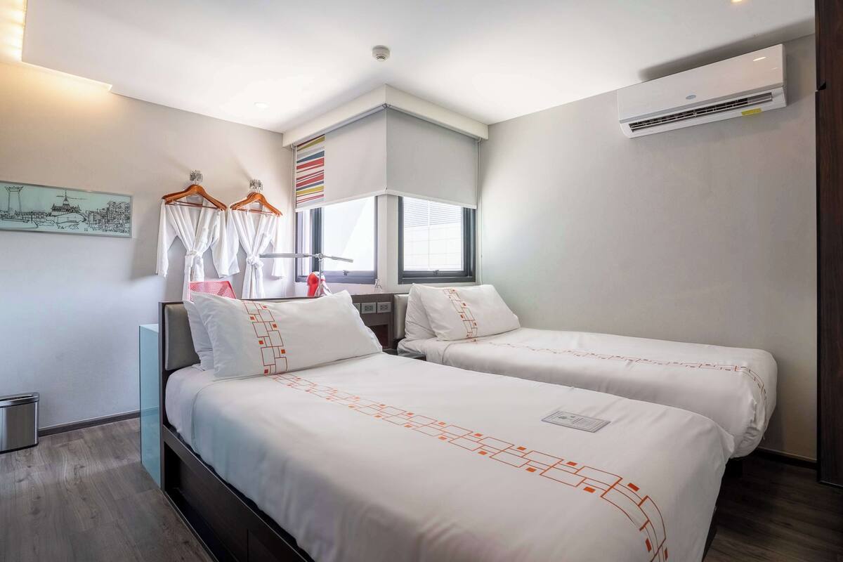 Une chambre d’hôtel avec deux lits, une fenêtre avec des stores, un appareil de climatisation et un tableau au mur.
