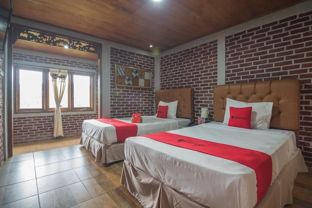 Quarto de hotel com duas camas, parede de tijolos, teto de madeira e janela com cortinas.