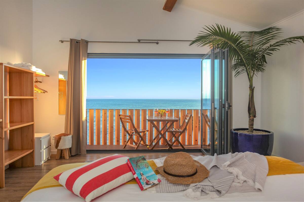 Un dormitorio con cama, armario de madera, pequeño taburete, mesa con sillas, planta en maceta y vista al mar a través de un ventanal.