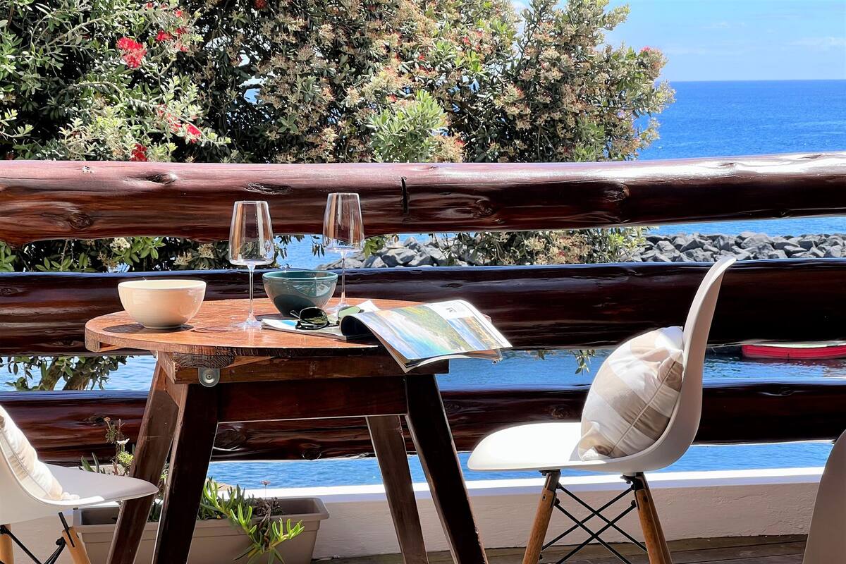 Una mesa de madera con un bol, dos copas de vino y una revista en un balcón con vista al mar.