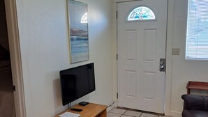 Un salon avec un téléviseur à écran plat posé sur un meuble en bois, une porte ornée d’un vitrail, un tapis à motifs et un canapé de couleur foncée.