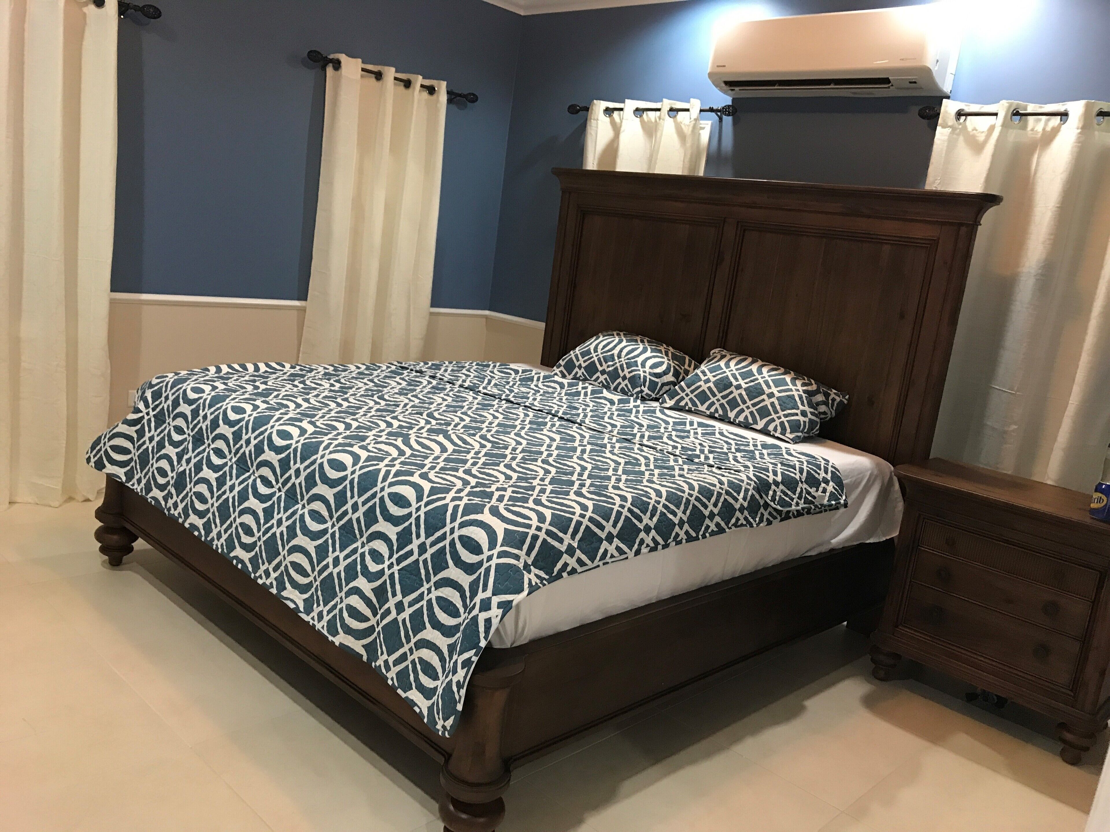 A bedroom with a wooden bed, white and blue patterned bedding, a wooden nightstand, and a ceiling air conditioner.