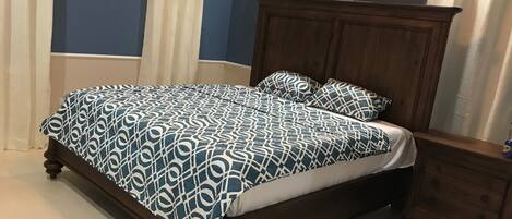 A bedroom with a wooden bed, white and blue patterned bedding, a wooden nightstand, and a ceiling air conditioner.