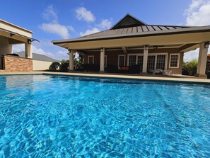 Une piscine à l’eau d’un bleu limpide, un patio couvert avec un toit et un mur en briques.